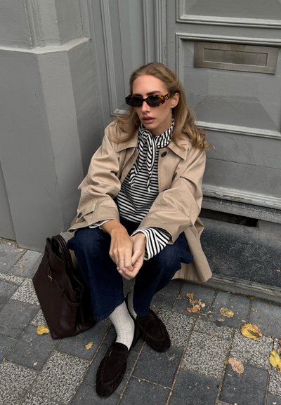 Trench beige, camiseta de rayas blanco y negro, jeans oscuros, bolso marrón, mocasines de ante oscuros, calcetines blancos, sentado en un pavimento de piedra estampado.