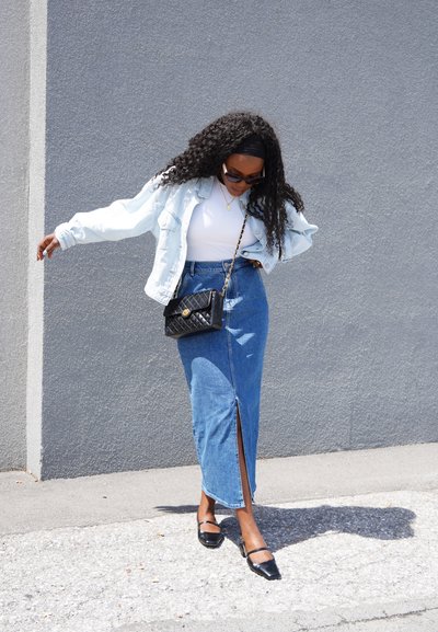 Chaqueta de mezclilla azul claro, blusa blanca ajustada, falda de mezclilla larga con una abertura frontal, bolso negro acolchado y zapatos negros con tiras.