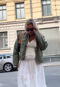 Žena v zelené bundě a bílé sukni si upravuje černé sluneční brýle na m�ěstské ulici s auty a budovami v pozadí.