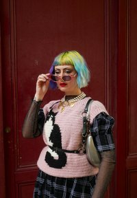 Ριχτό ροζ πουλόβερ πλεξίματος με σχέδιο μαύρης γάτας, τοποθετ ημένο πάνω από ένα καρό φόρεμα σε ασπρόμαυρους τόνους; διαφανή γυαλιά ηλίου; ασημένια τσάντα; πολύχρωμα κοντά μαλλιά.