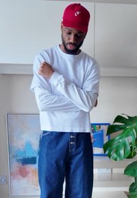 Wit shirt met lange mouwen gecombineerd met blauwe high-waisted denim jeans met twee gouden knopen. Rode pet met logo aan de voorkant.