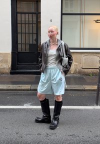Schwarze Lederstiefel bis zum Knie, hellblaue Shorts mit seitlichen Streifen, grauer Kapuzenpullover unter einer braunen Lederjacke und eine kleine helle Tasche.
