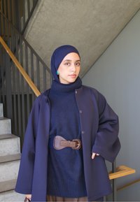 Palton supradimensionat bleumarin suprapus peste un pulover cu guler înalt asortat, strâns cu o curea maro. Purtați cu un hijab albastru și cercei turcoaz.