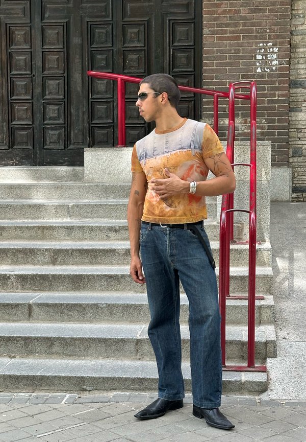 Mann mit Sonnenbrille, bedrucktem T-Shirt und Jeans steht auf Steinstufen neben einem roten Geländer und blickt zur Seite vor einer dunklen Holztür.