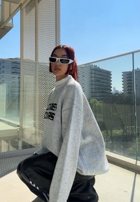 Suéter de punto en gris claro con texto oscuro, diseño oversize. Pantalones negros y brillantes y gafas de sol blancas con forma angular. Entorno exterior brillante.