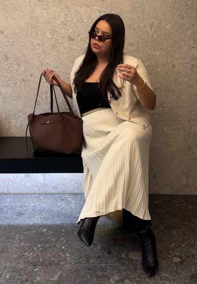 Mujer con cabello largo y oscuro que lleva gafas de sol, un cárdigan crema abotonado, camiseta negra, falda plisada crema y botas negras, sosteniendo un bolso grande marrón.