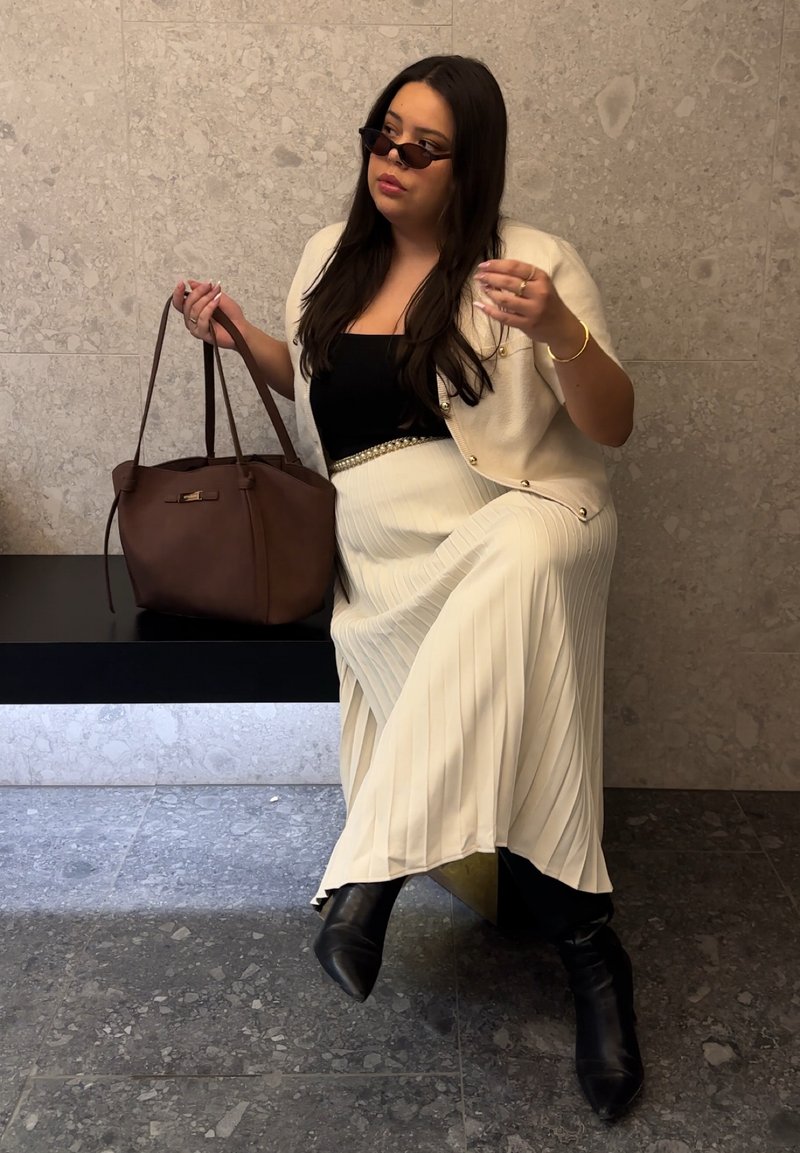 Femme aux longs cheveux noirs portant des lunettes de soleil, un cardigan crème boutonné, un haut noir, une jupe plissée crème et des bottes noires, tenant un grand sac à main marron.