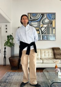 Chemise à rayures bleu clair, pantalon beige, pull noir noué à la taille et chaussures noires. Cadre intérieur avec un canapé blanc et une œuvre d'art en arrière-plan.