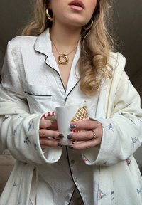 Haut de pyjama blanc à pois avec passepoil noir, cardigan en tricot crème avec motifs, tenant une tasse à motif ; accessoires comprenant des boucles d'oreilles créoles dorées et un collier chaîne.
