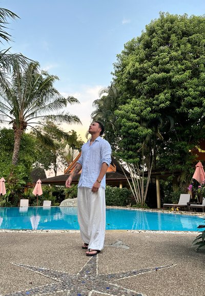 Hombre con una camisa de manga larga azul claro y pantalones blancos sueltos, de pie cerca de una piscina con palmeras y sombrillas rosas al fondo.
