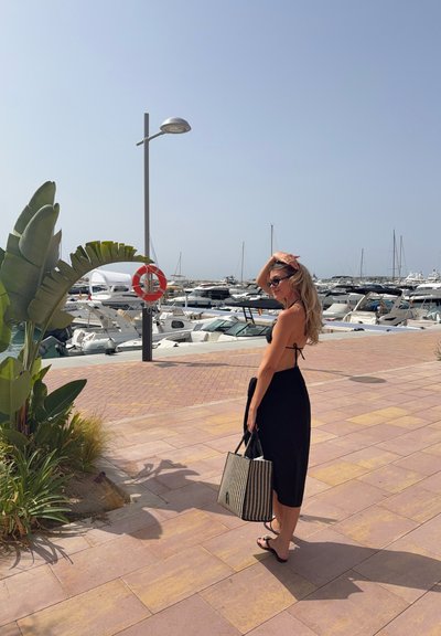Vestido negro con tirantes finos, llevando un bolso de rayas, caminando por un sendero pavimentado cerca de una marina llena de barcos y vegetación.