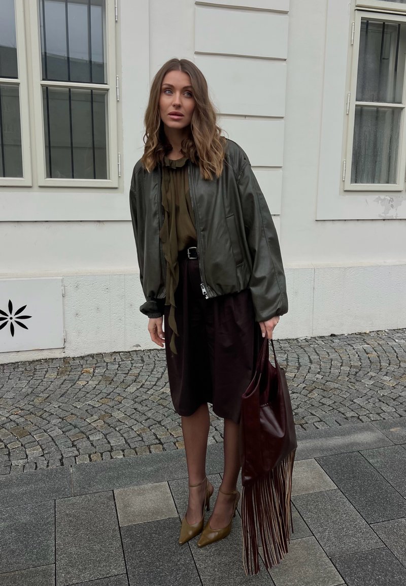 Frau mit welligem Haar, die eine übergroße dunkelgrüne Jacke, schwarzen Rock und olivfarbene Absatzschuhe trägt, hält eine große braune Fransen-Tasche und steht auf einer Kopfsteinpflasterstraße.