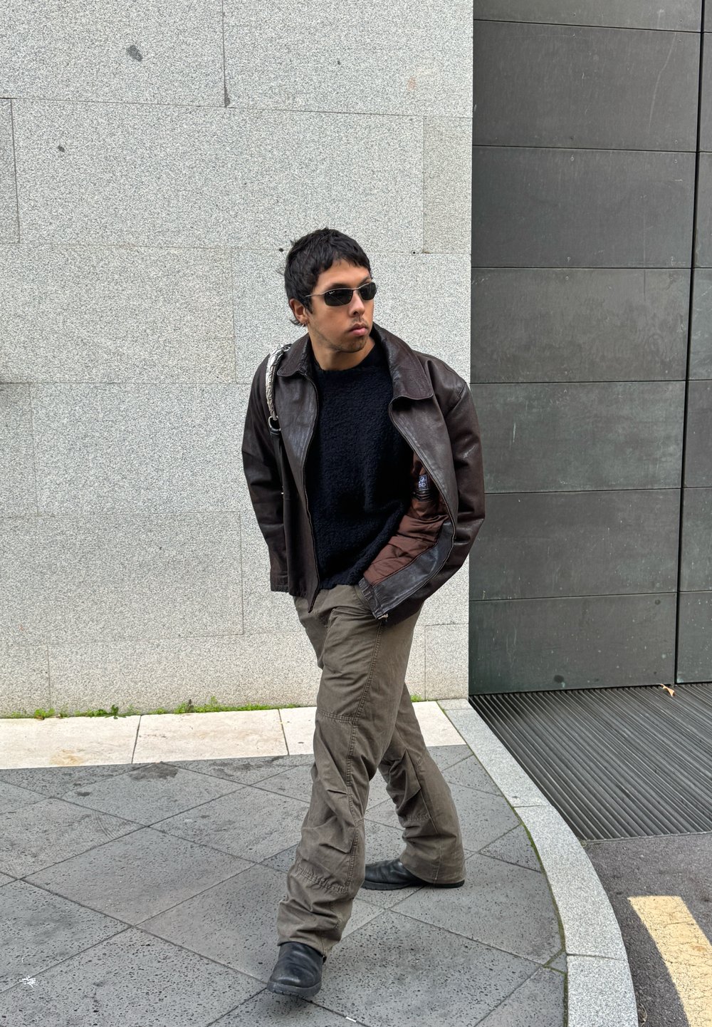 Καφέ δερμάτινο μπουφάν, μαύρος υφασμάτινος φούτερ, ελαιοπράσινα cargo παντελόνια, μαύρες μπότες και γυαλιά ηλίου, περπατούν μπροστά σε γκρι πέτρινο φόντο.