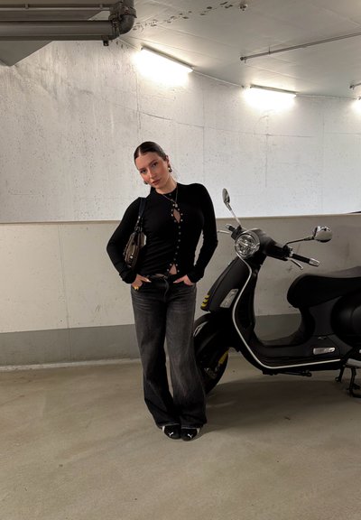 Joven con cabello oscuro, vestida con una blusa negra y jeans, está de pie junto a un scooter negro en un estacionamiento de concreto.