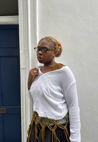 Weißes, geripptes Langarmshirt, darüber ein gelb-grün gemusterter Träger. Schwarzer Rock mit geknoteten Senfakzenten, vor einer hellen Wand.