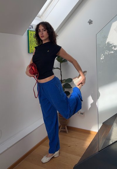 Mujer con cabello rizado, vestida con top negro y pantalones azules, sosteniendo un bolso rojo y estirando una pierna hacia atrás en un espacio interior luminoso.