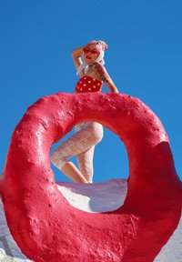 Rdeče bodičasto kopalke in bele čipkaste pajkice na modelu, ki je postavljen na vrhu velike rožnate okrogle skulpture pod jasnim modrim nebom.