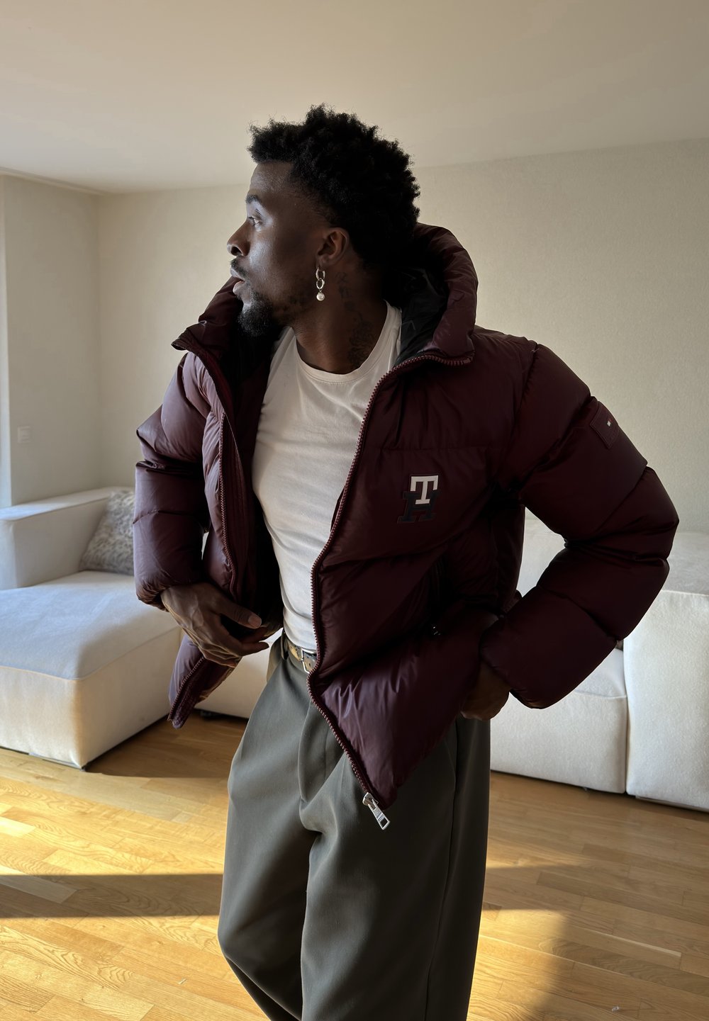 Homme aux cheveux courts et bouclés, portant une boucle d'oreille, vêtu d'une doudoune bordeaux par-dessus une chemise blanche et un pantalon gris ample dans une pièce lumineuse.