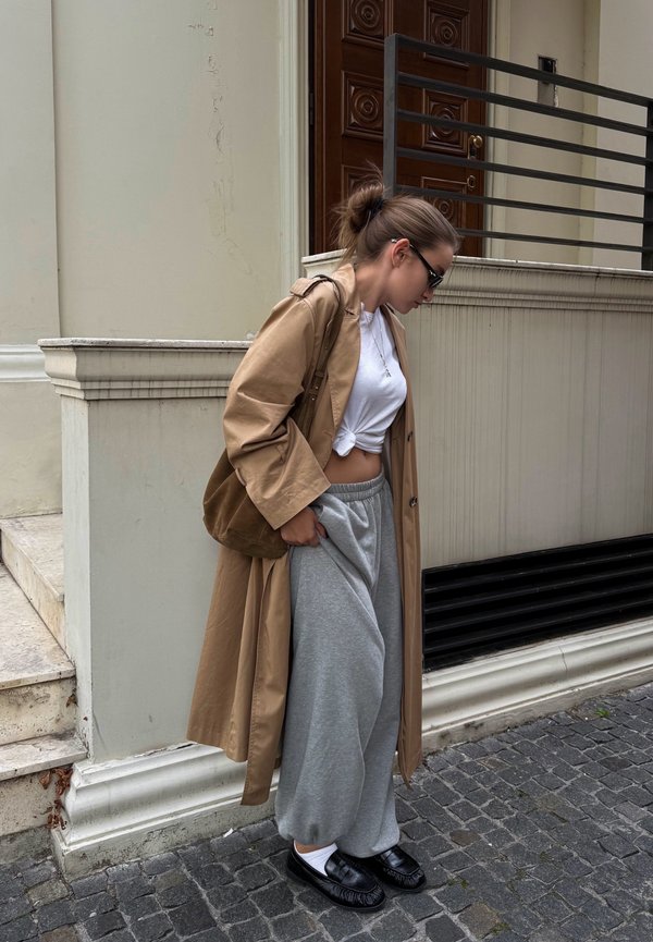Femme portant un trench-coat beige, une chemise blanche nouée, un pantalon de survêtement gris, des chaussures noires et des lunettes de soleil marchant dans une rue pavée.
