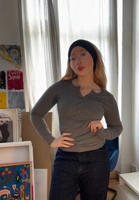 Pelēka garām piedurknēm henley krekls ar noapaļotu apkakli, saskaņots ar tumši ziliem džinsiem. Fons ietver spilgti krāsotus mākslas darbus.