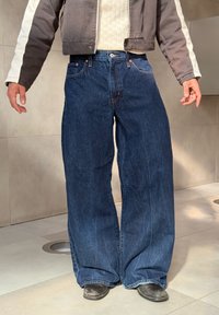 Bredde bukser i mørkeblå denim med kontrastsying, forlommer og en afslappet pasform. Iført en grå jakke.