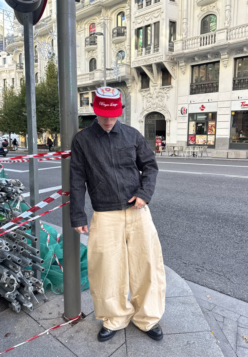 Fekete csíkos dzseki cipzáras előlappal és egy zsebbel, széles bézs nadrággal és piros baseball sapkával, amelyen grafikás mintázat látható.