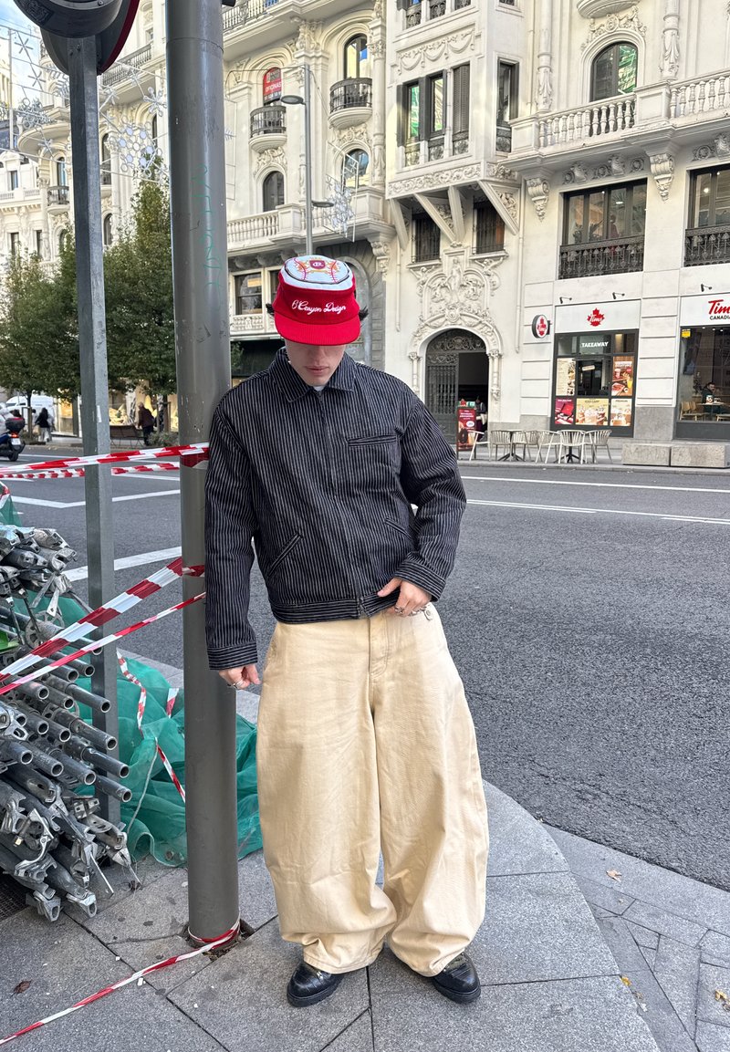 Black striped jacket with a zip front and one pocket, paired with wide beige trousers and a red baseball cap with a graphic design.