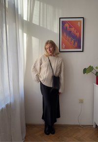 Moteris, vilkinti kreminės spalvos megztiniu su pynėmis ir juoda sijonu, pozavo saulėtoje patalpoje su rėmintu abstrakčiu menu ir vazonine planta ant baltų baldų.