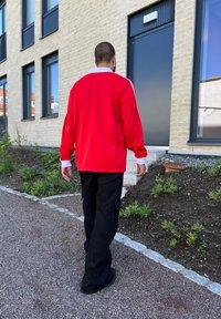 Casaco vermelho com fecho éclair e detalhes brancos, combinado com calças cargo pretas, a caminhar por um caminho de gravilha do lado de fora de um edifício moderno.