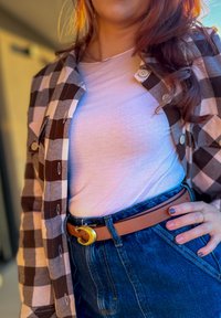 Hnědo-bílá kostkovaná flanelová košile přes bílé tričko, doplněná vysokým modrým džínsem a hnědým páskem se zlatou přezkou.