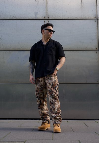 Camisa corta de manga negra con textura, pantalones cargo con estampado floral y botas marrones. Los accesorios incluyen gafas de sol y un reloj.