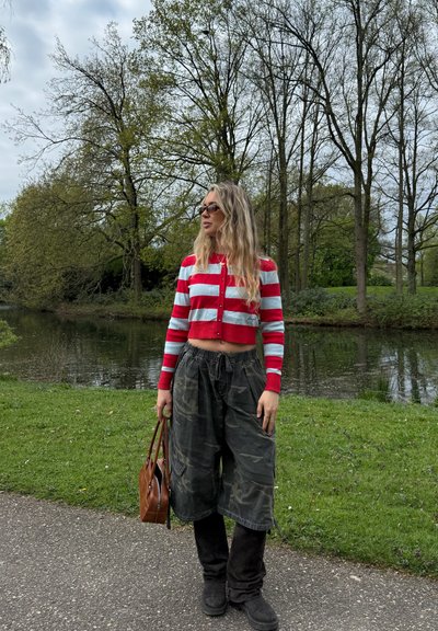 Mujer con cabello largo y rubio y gafas de sol, de pie cerca de un estanque, lleva un suéter corto de rayas rojas y blancas, pantalones holgados de camuflaje y botas negras.