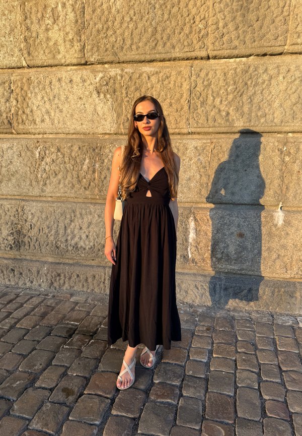 Una mujer con gafas de sol y un vestido negro está de pie sobre un adoquinado junto a un muro de piedra iluminado por el sol, proyectando una larga sombra.