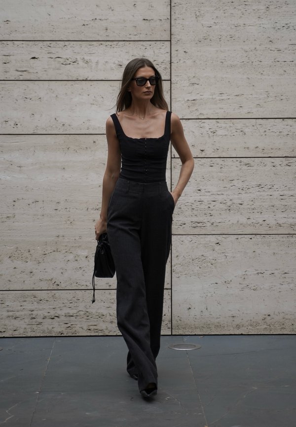 Woman walking in black sleeveless top and wide-leg pants, carrying a small black bag, with sunglasses against a stone wall background.