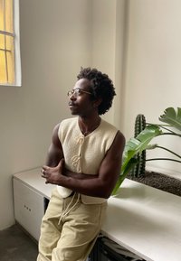 Gilet beige sans manches en tricot avec fermeture à lacet, porté sur un pantalon kaki clair. En arrière-plan, un cactus en pot et de larges feuilles vertes.