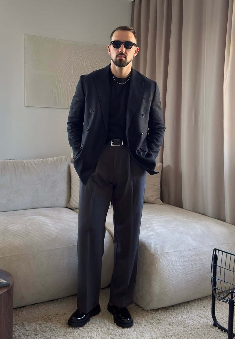 Homme portant des lunettes de soleil noires, un blazer noir croisé, une chemise noire, un pantalon gris et des chaussures noires, debout dans un salon.