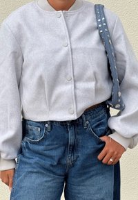 Giacca bomber in pile grigia con chiusura a bottoni, polsini a costine, abbinata a jeans blu in denim a vita alta e una tracolla grigia con borchie.