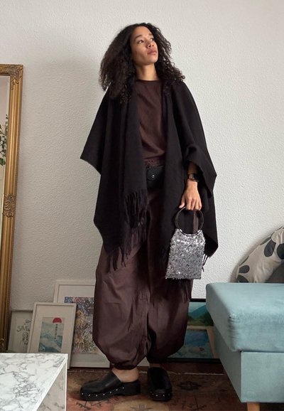 Mujer con cabello rizado está dentro de un edificio vistiendo ropa suelta de color marrón oscuro y un chal negro, sosteniendo un pequeño bolso plateado con lentejuelas.