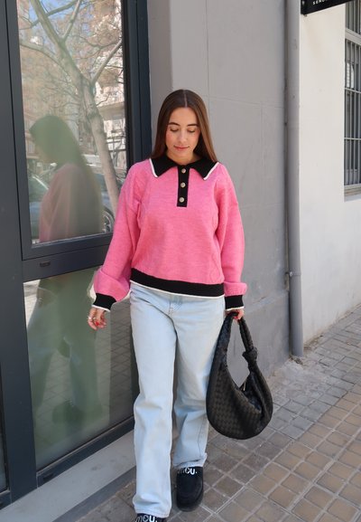 Jersey rosa con cuello y botones negros, combinado con jeans de un azul claro y bolso negro. Estilo casual con tejido texturizado y detalles en contraste.