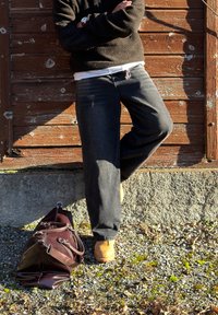 Personne appuyée contre un mur en bois usé, bras croisés, portant un pull noir, un jean gris, des bottes couleur sable et un sac de voyage en cuir marron posé au sol.