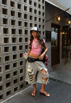 Joven con shorts grises rotos, top corto rosa y gorra, de pie sosteniendo una bebida frente a una pared de concreto con patrón, cerca de la entrada de un café.