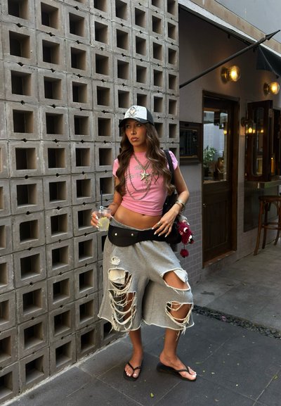 Joven con shorts grises rotos, top corto rosa y gorra, de pie sosteniendo una bebida frente a una pared de concreto con patrón, cerca de la entrada de un café.