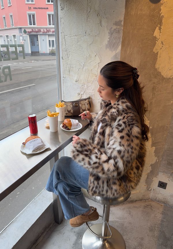 Frau in einem Leopardenmuster-Mantel sitzt auf einem Hocker am Fenster und isst Burger und Pommes, mit einer Getränkedose und einer Handtasche auf dem Tisch.