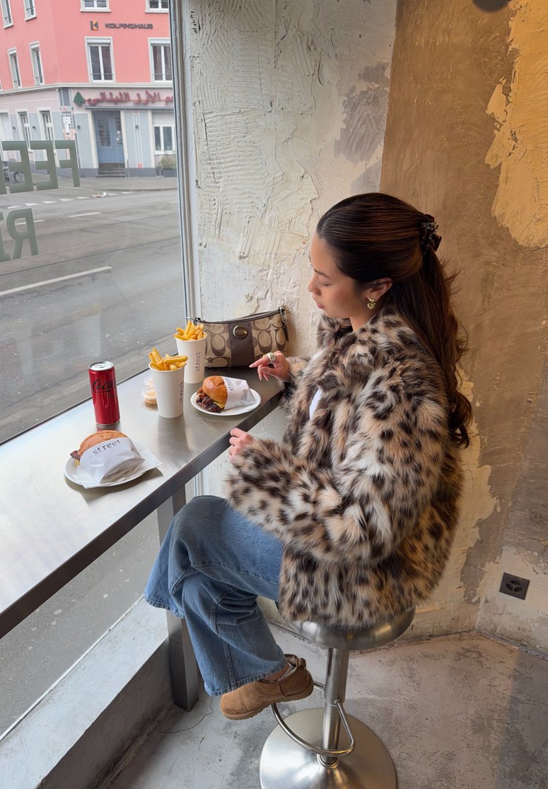 Kvinde iført leopardmønstret frakke, der sidder på en skammel ved et vindue og spiser burger og pommes frites, med en dåse drik og en håndtaske på bordet.