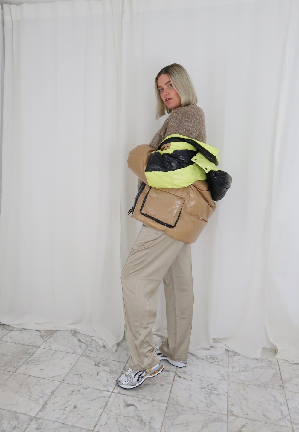 Casaco puffer bege com detalhes em preto e amarelo fluorescente, suéter creme, calças bege e tênis de corrida prateados num chão de mármore.