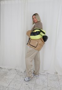 Casaco puffer bege com detalhes em preto e amarelo fluorescente, suéter creme, calças bege e tênis de corrida prateados num chão de mármore.