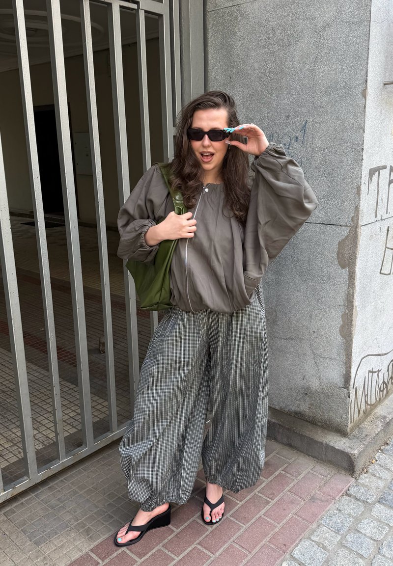 Femme portant des lunettes de soleil, une veste grise oversize, un pantalon large à carreaux, des sandales compensées noires et portant un sac vert, debout près d'un portail métallique et d'un mur en béton.