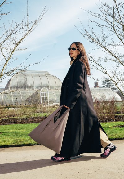 Una mujer que lleva un abrigo negro y gafas de sol carga una bolsa tote beige y usa calzado negro con acentos rosas, caminando en un jardín.