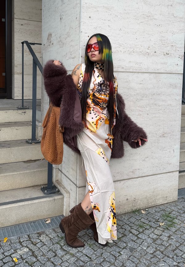 Personne aux longs cheveux noirs et colorés, portant des lunettes rouges, une robe à motifs, une veste en fourrure marron et des bottes, s'appuie contre un mur de pierre clair près des marches.