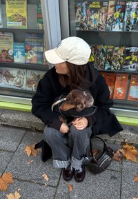 Oseba prikrčena, obleka črni plašč in bež kapa. Drži rjavega psa v črni prešiti jakni. Temno modre kavbojke in sijoči čevlji. Blizu je rjava torba.